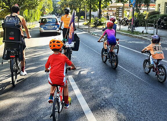 Kidical Mass Schöneberg September 2024 Eine Gruppe Kinder fährt bei sonnigem Wetter auf Fahrrädern das Berliner Reichpietschufer entlang, auf der Fahrbahn. Vorne fährt ein Auto der Polizei Berlin als Führungsfahrzeug, und zwei Erwachsene. Es ist eine ADFC Berlin Fahne zu sehen.